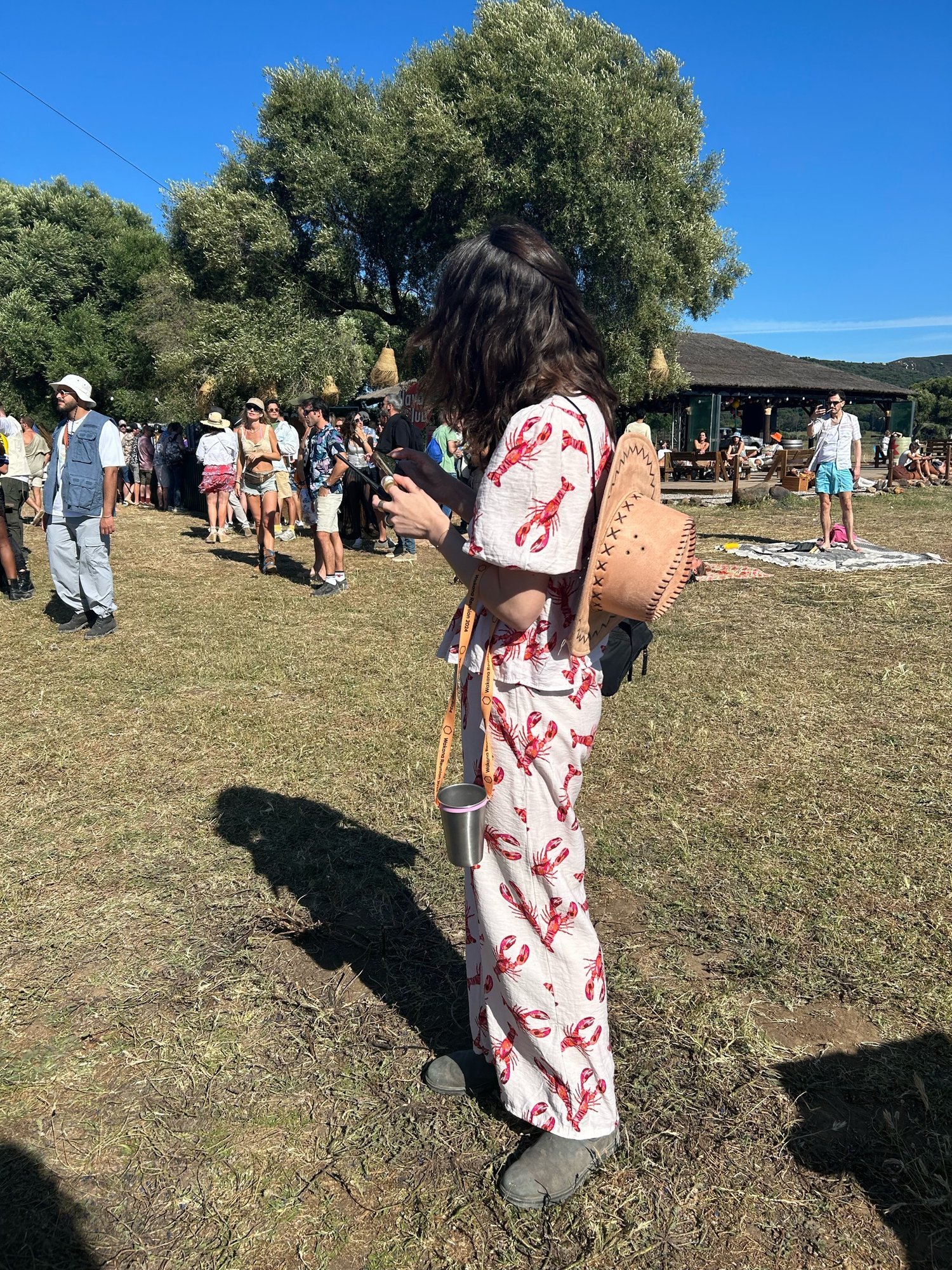 Mujer de pie en un festival al aire libre, vestida con un conjunto de pantalón y blusa blanca con estampado de langostas rojas. Lleva un sombrero de paja y sostiene un vaso metálico con una correa. A su alrededor, se ven más personas disfrutando del evento en un día soleado.