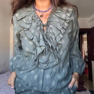 Persona usando una blusa verde con volantes alrededor del cuello y pecho, con un patrón de pequeños diamantes estampados. Lleva un collar azul con un dije en forma de flor.