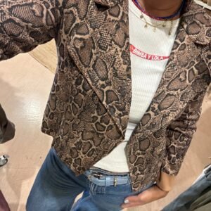 Persona usando una chaqueta de estampado de serpiente en tonos marrones, sobre una camiseta blanca con texto rojo. Lleva jeans de cintura alta y un cinturón azul claro.