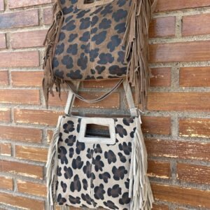 Dos bolsos de gamuza con estampado de leopardo, uno marrón oscuro y otro beige, colgados sobre una pared de ladrillo. Ambos tienen flecos largos a los lados y asas rectangulares.