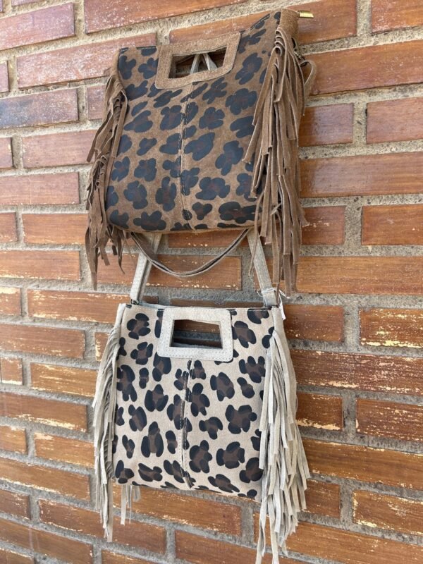 Dos bolsos de gamuza con estampado de leopardo, uno marrón oscuro y otro beige, colgados sobre una pared de ladrillo. Ambos tienen flecos largos a los lados y asas rectangulares.