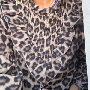 Persona vistiendo una blusa con estampado de leopardo en tonos marrón y negro, con detalles fruncidos y volantes en el escote. Lleva un collar de cuentas azules.