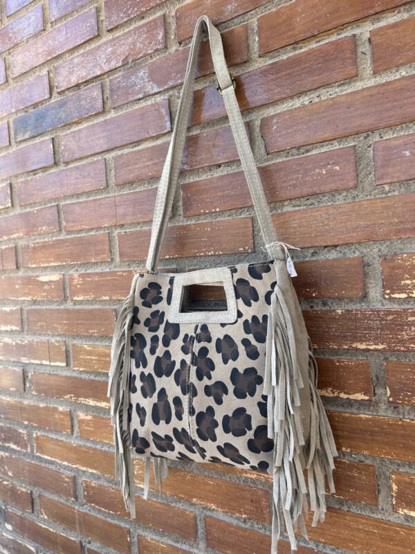 Bolso beige con estampado de manchas negras y flecos, colgado sobre una pared de ladrillos.