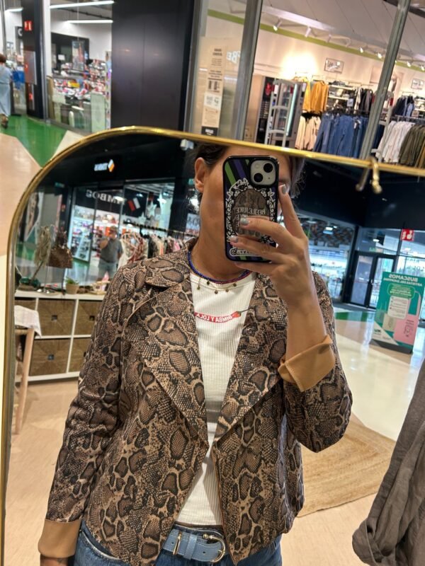 Persona tomándose una foto en un espejo, usando una chaqueta con estampado de serpiente en tonos marrón, sobre una camiseta blanca con texto rojo.