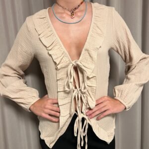 Blusa beige con lazos ajustables en la parte delantera y volantes en el escote, usada con pantalones oscuros.