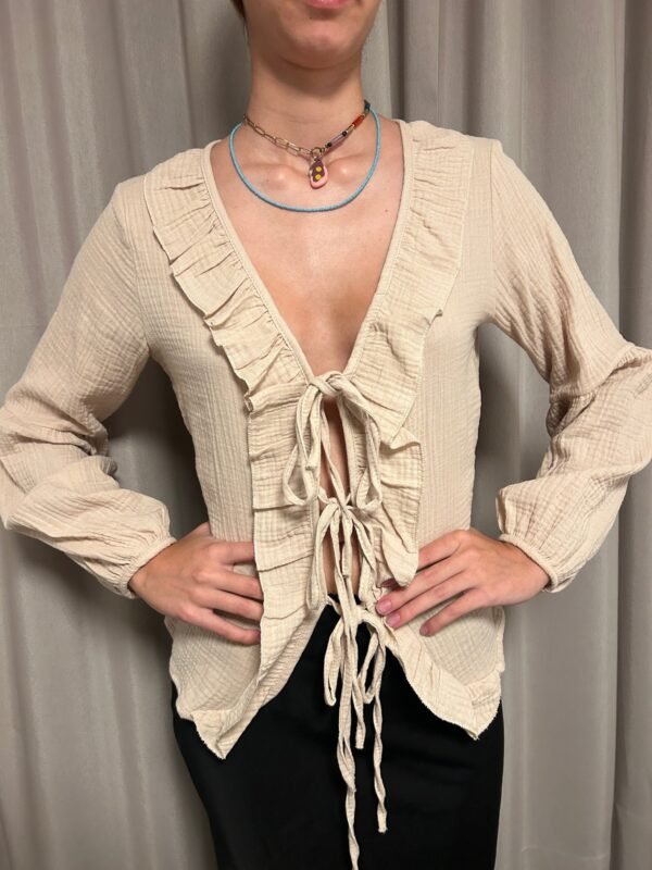 Blusa beige con lazos ajustables en la parte delantera y volantes en el escote, usada con pantalones oscuros.