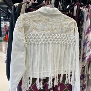 Chaqueta blanca colgada en un perchero, con bordados florales en hilo beige y un diseño de flecos largos que cuelgan desde la parte trasera.